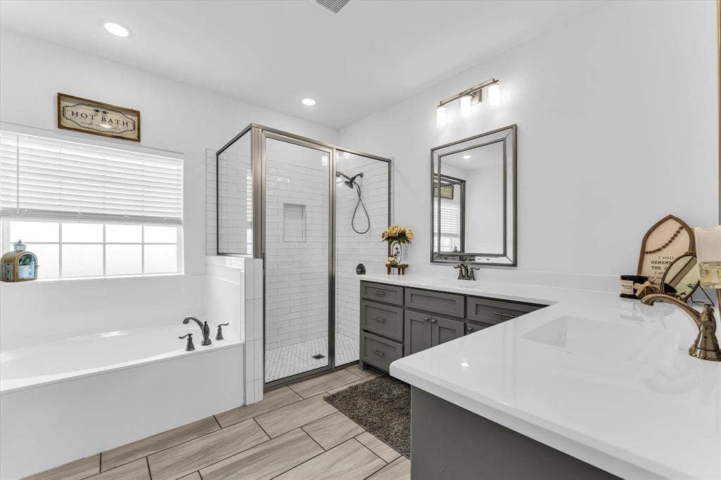 Spacious bathroom featuring a built-in soaking tub, glass-enclosed shower with subway tile, wood-finish tile flooring, and a double vanity with grey cabinetry and white countertops