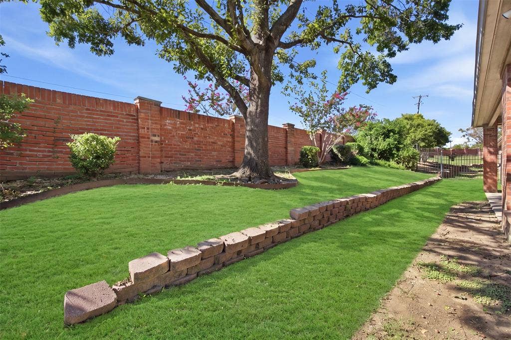 750 Village Green Drive, DeSoto, TX 75115 Green lawn with mature shade trees, bordered by a brick privacy wall and a stone retaining wall. Green enhancement to lawn.