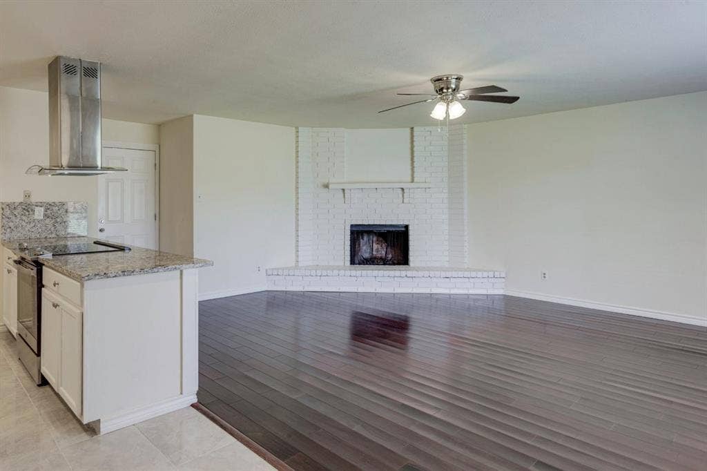 Wood-finish flooring, white painted brick fireplace with raised hearth and mantel, stainless steel chimney-style range hood, granite-topped kitchen island, and ceiling fan