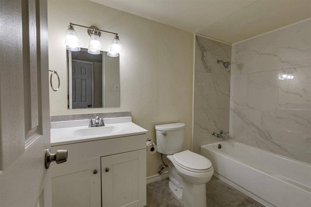 Bathroom featuring a white vanity cabinet with a solid-surface countertop, an integrated sink, three-light vanity fixture, and a shower-tub combination with full-height stone-finish wall tile