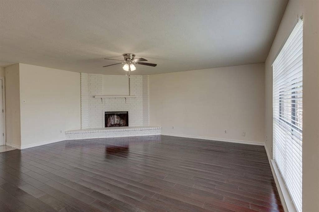 Spacious interior featuring wood-finish flooring, a corner fireplace with a brick surround, a mantel, and a ceiling fan with integrated lighting