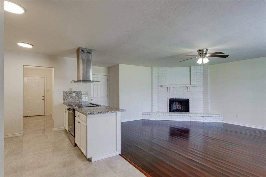White-painted brick fireplace with a wood mantel, stainless steel island-mounted range hood, granite countertops, wood-finish flooring, and recessed ceiling lighting