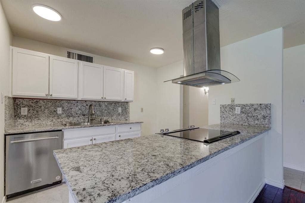 Kitchen featuring speckled stone countertops, white shaker-style cabinetry, stainless steel dishwasher, glass-accented stainless steel range hood, and a flat-surface cooktop