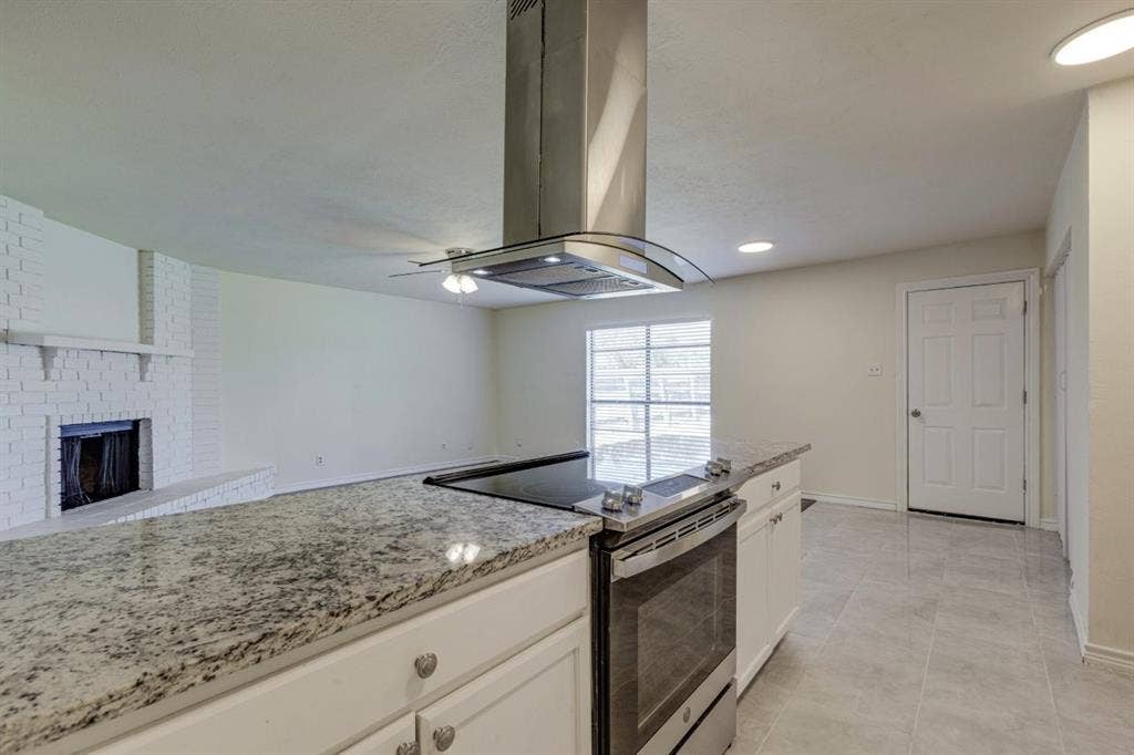 Kitchen featuring a stone-surface island, stainless steel range with overhead chimney vent, white cabinetry, tile flooring, and a painted brick fireplace