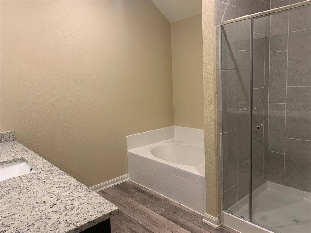 Built-in soaking tub, walk-in shower with glass enclosure, wood-finish flooring, and white speckled countertop