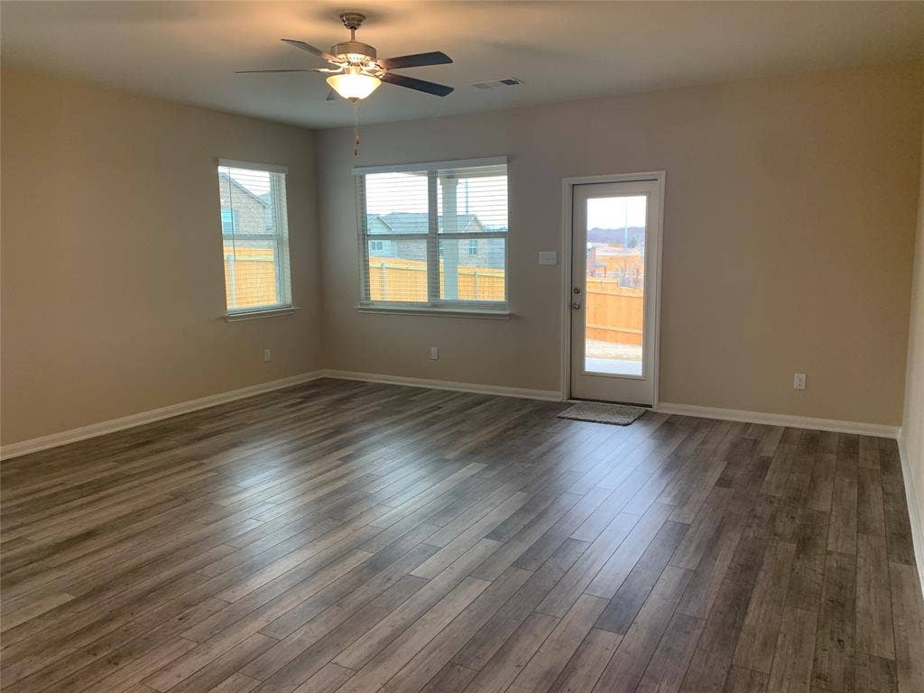 Expansive room featuring wood-finish flooring, neutral-toned walls, a ceiling fan with integrated lighting, multiple windows, and a glass-paneled exterior door