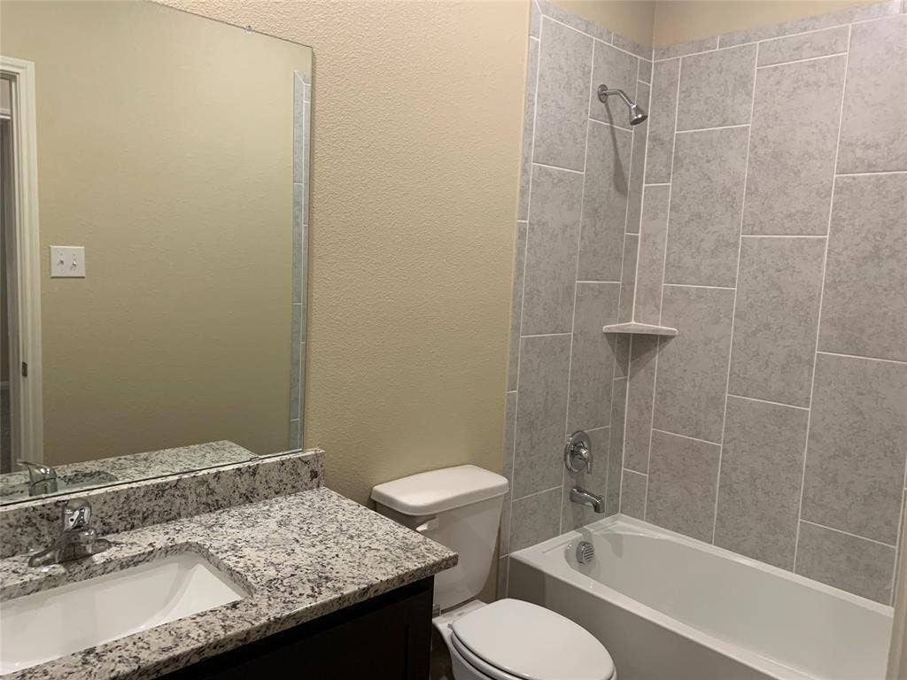 Vanity with built-in sink and stone-finish countertop, mirror with decorative trim, and shower-tub combination with tiled surround and built-in shelf