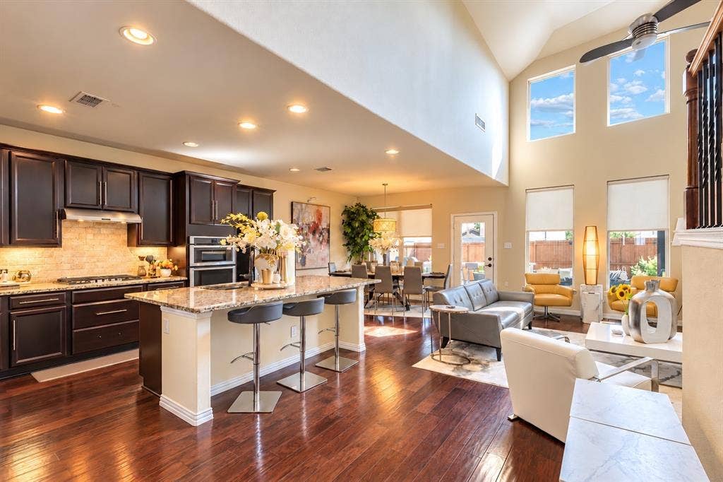 1704 Keswick Drive, McKinney, TX 75071 Open-concept kitchen and living area featuring dark cabinetry with stainless steel appliances, granite countertops, a large center island with seating, hardwood flooring throughout, and floor-to-ceiling windows with abundant natural light and skylight accents