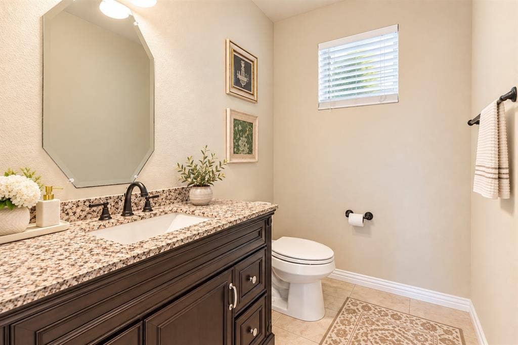1704 Keswick Drive, McKinney, TX 75071 Elegant bathroom featuring a dark wood vanity with dual-sink functionality, granite-look countertop, octagonal framed mirror with integrated lighting, and a spacious layout