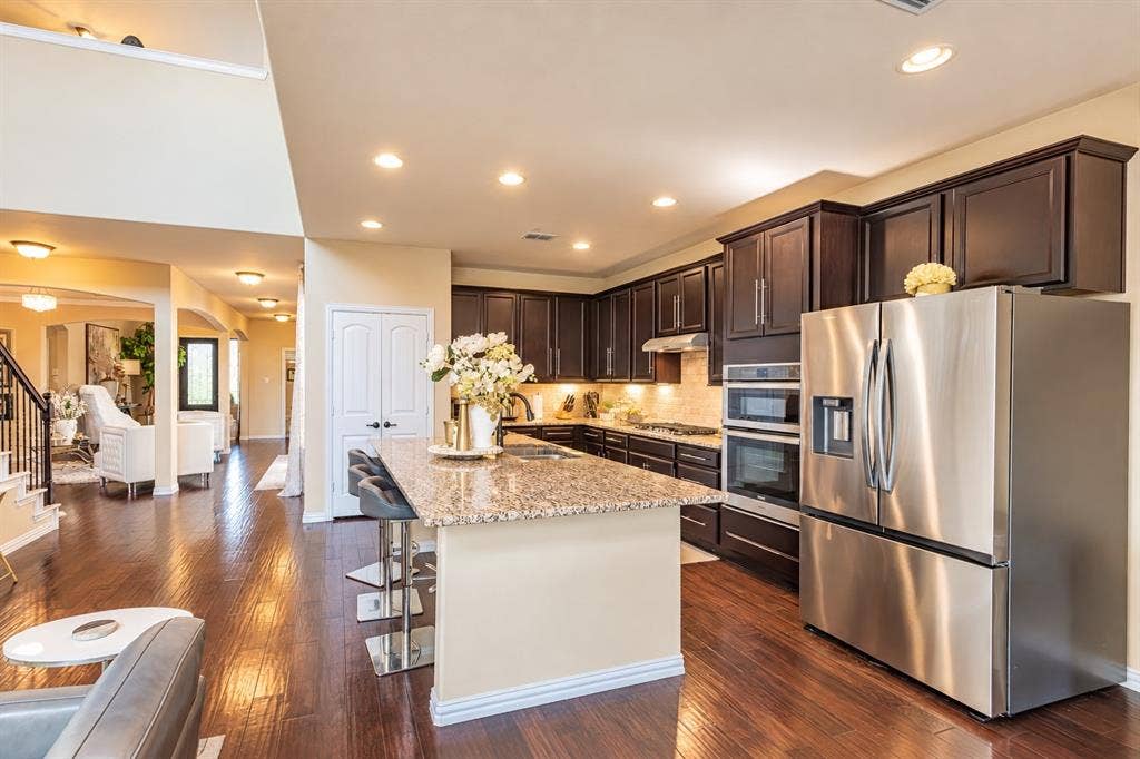 1704 Keswick Drive, McKinney, TX 75071 Chef's kitchen featuring dark cabinetry, stainless steel appliances including dual wall ovens, granite countertops, a large center island with seating, recessed lighting, and an open layout connecting to the main living spaces