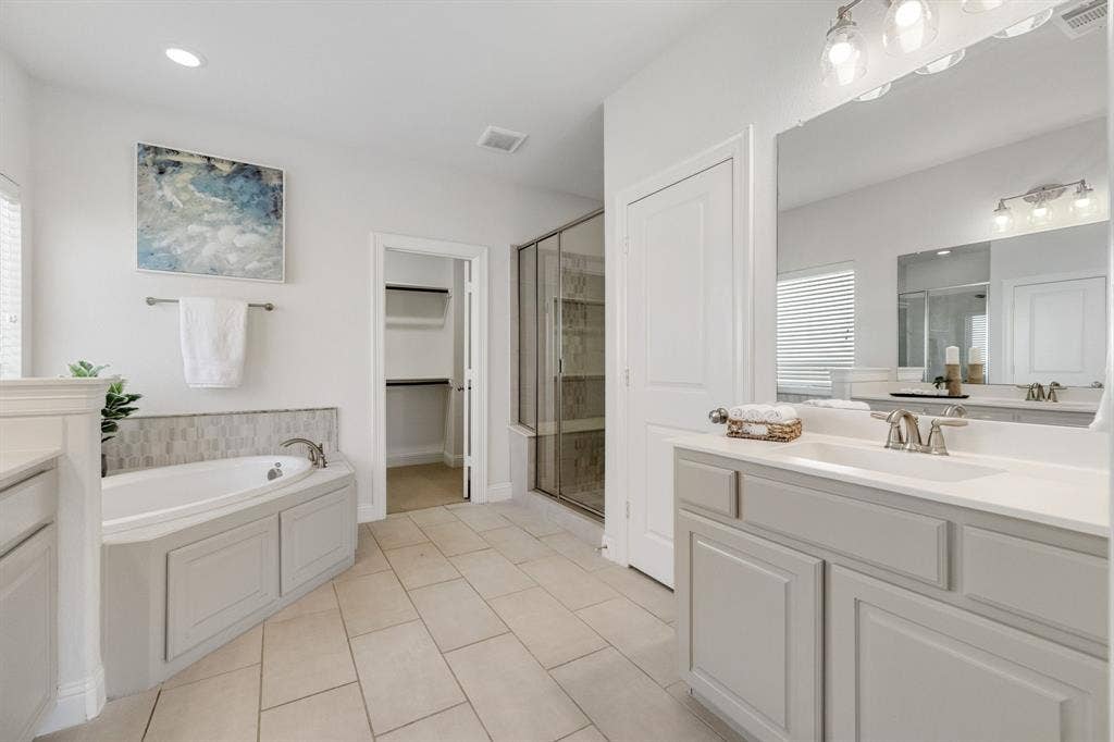 Spacious bathroom with neutral tile flooring, a built-in corner soaking tub, a separate glass-enclosed shower, a large vanity with an integrated sink, and an expansive wall-mounted mirror