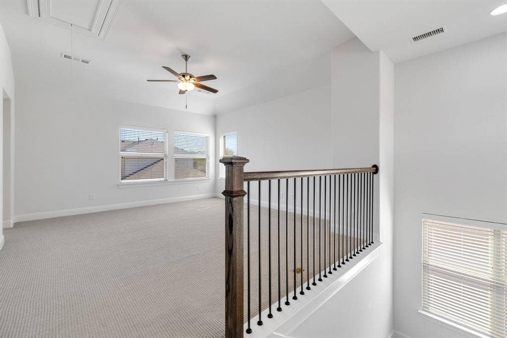 Spacious second-level area featuring light-colored carpeting, multiple windows with blinds, a ceiling fan with integrated lighting, and a stair railing with wood newel posts and black metal balusters