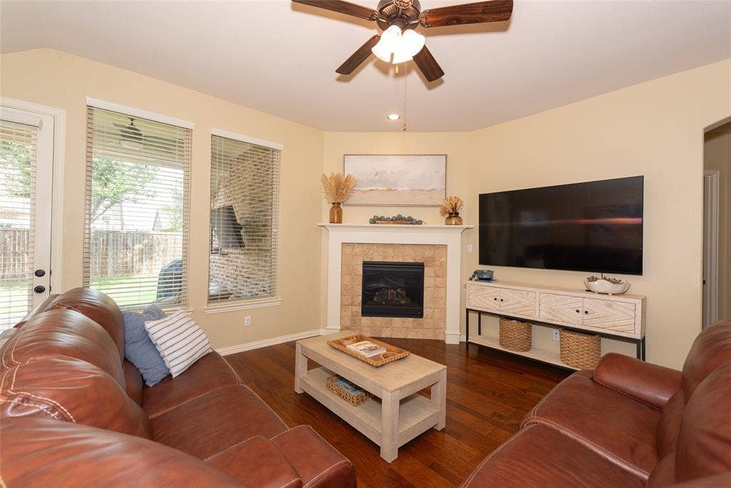 Wood-finish flooring, corner fireplace with tile surround, ceiling fan, and multiple windows