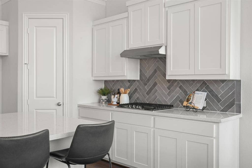 1320 Cartona Road, Fate, TX 75087 White shaker cabinetry, light-toned countertops, herringbone tile backsplash, stainless steel range hood, and a built-in gas cooktop