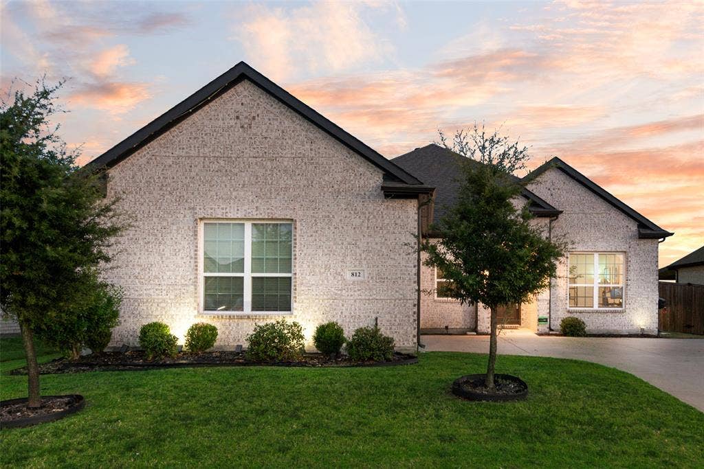 812 Stamford Avenue, Arlington, TX 76002 White brick exterior, black shingle roof, integrated landscaping with shrubs and trees, manicured lawn, and paved driveway
