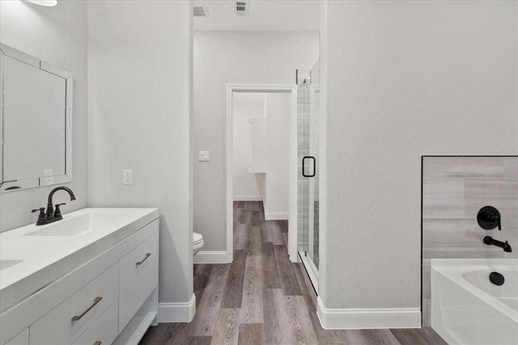 7919 N kiska Street, Dallas, TX 75216 White vanity with matte black faucet, framed mirror, glass-enclosed shower, white bathtub with black fixtures, and wood-finish flooring