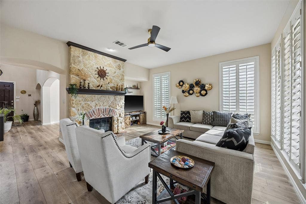 2865 Ridge View Road, Frisco, TX 75034 Stone-clad fireplace with a brick arch and dark wood mantel, wood-finish flooring, white plantation shutters, and a ceiling fan with integrated lighting