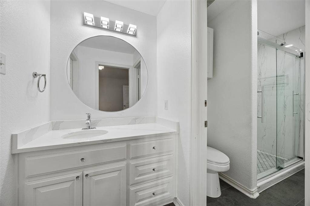 2865 Ridge View Road, Frisco, TX 75034 White vanity with integrated sink and chrome faucet, large circular mirror, four-light vanity fixture, glass-enclosed shower with subway tile, and dark floor tiling
