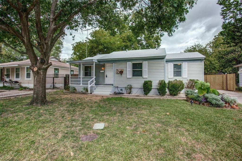 Single-story residence featuring light-colored siding, a covered porch with white railings, and a landscaped front yard with established plantings