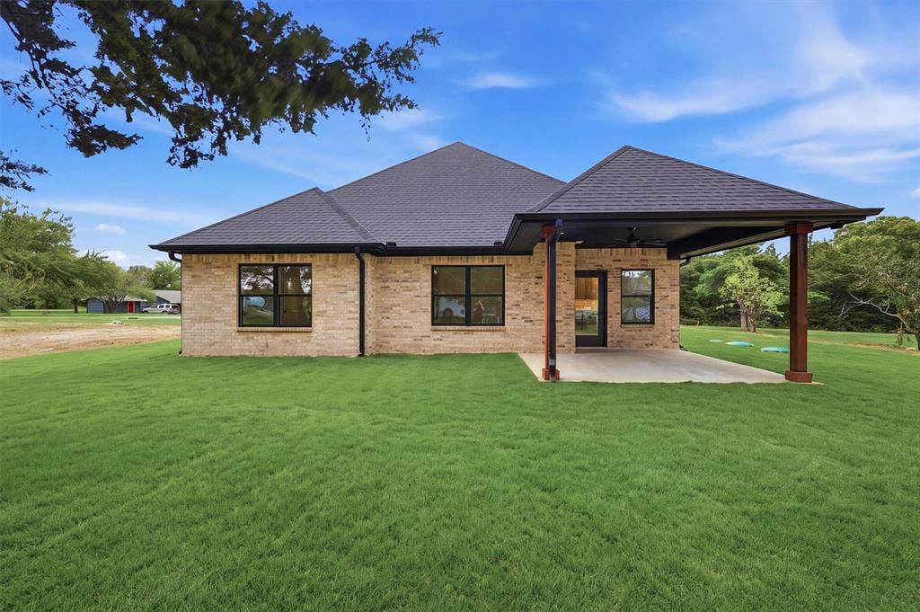 Light brick exterior featuring a covered patio, dark-toned architectural roof shingles, black-framed windows, and a expansive manicured lawn