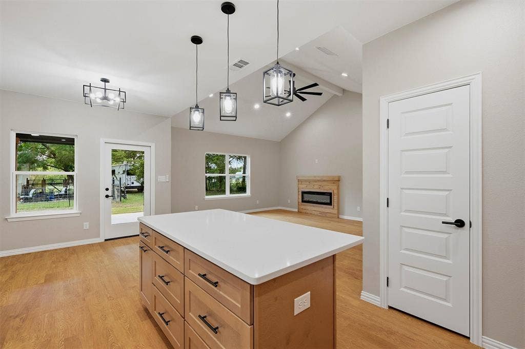 Open-concept interior featuring vaulted ceilings, a kitchen island with stone countertop, wood-finish flooring, matte black pendant lighting, and a fireplace with a wood mantel