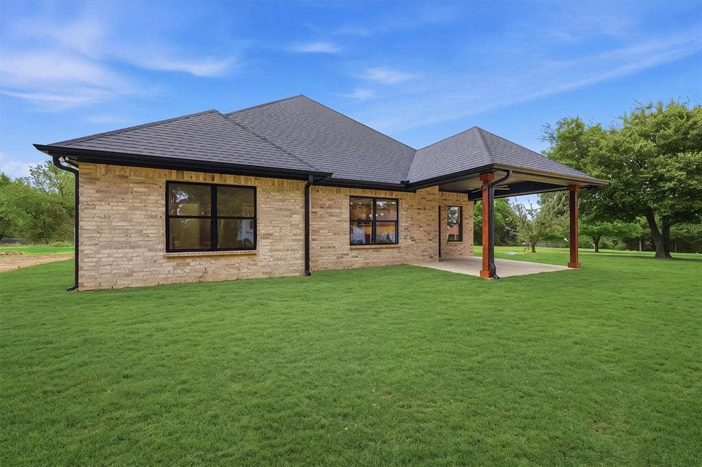 Exterior featuring light-toned brick masonry, a covered rear patio with wood-finish structural support posts, dark-framed windows, and a multi-pitched roof with charcoal-toned shingles
