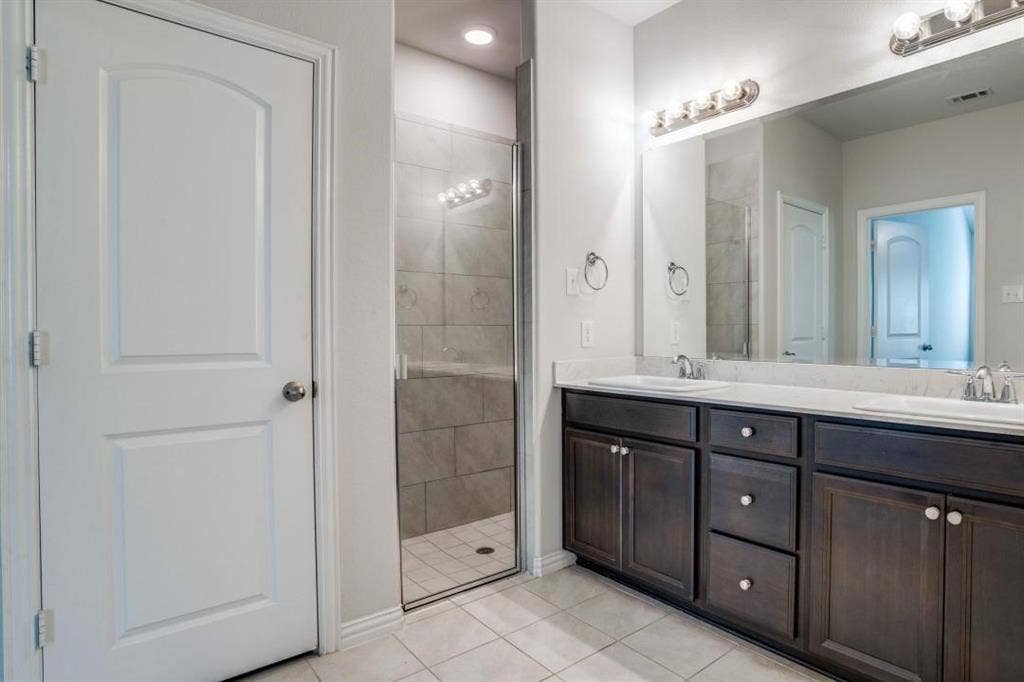 6229 Gloucester Drive, Celina, TX 76227 Dual vanity with dark wood-finish cabinetry and white countertops, large mirror, and glass-enclosed shower with tiled walls