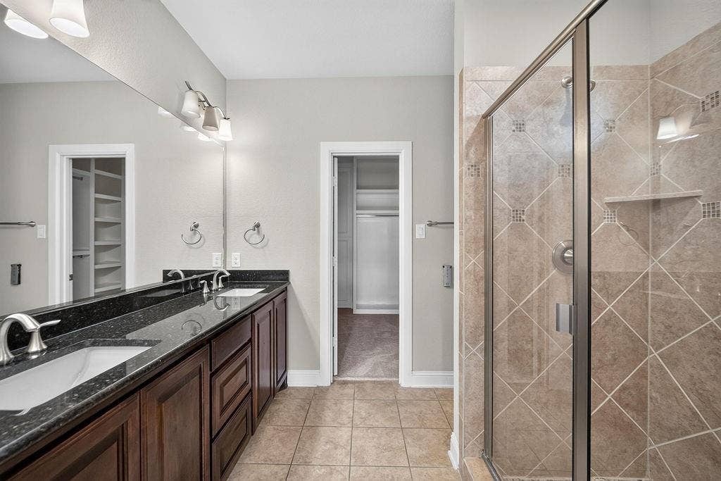 Dual-sink vanity with dark stone countertop, wood cabinetry, full-width mirror, and walk-in shower with diamond-pattern tile surround