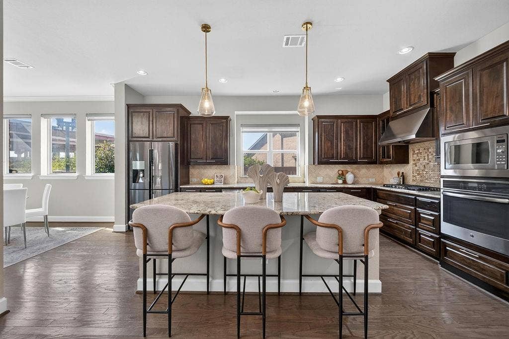 Kitchen featuring dark wood cabinetry, an expansive center island with a stone countertop, stainless steel built-in appliances, a tiled backsplash, and wood-finish flooring