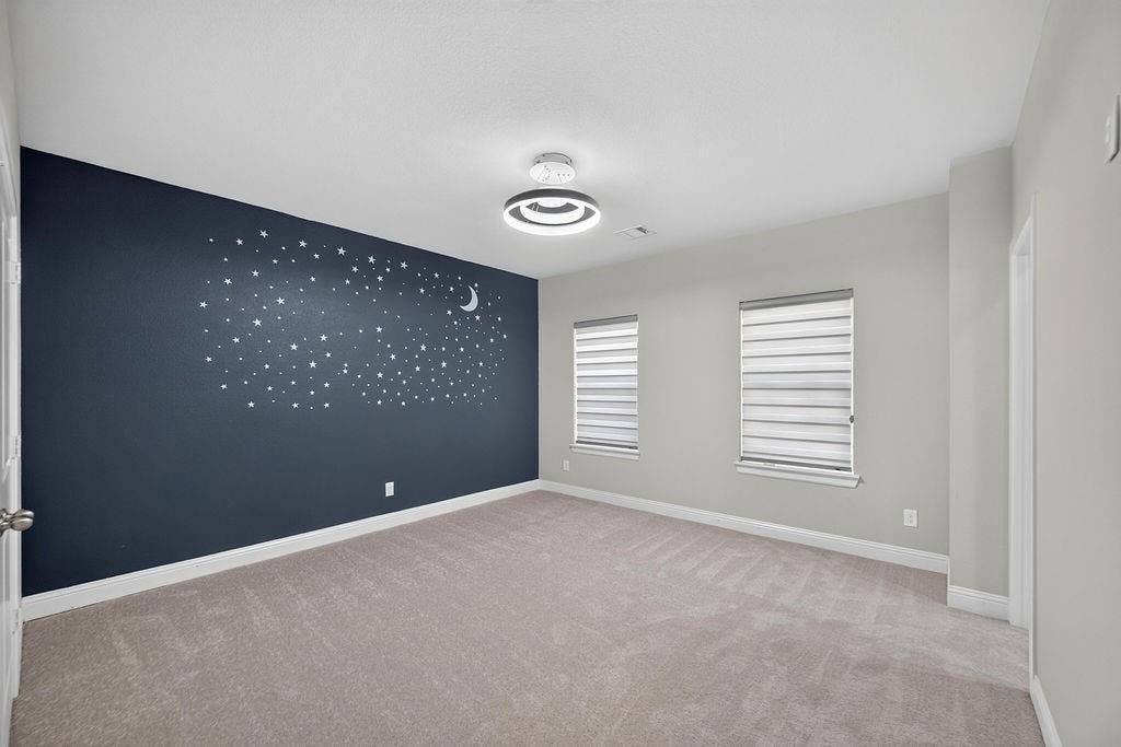 Neutral-toned room featuring wall-to-wall carpeting, a dark accent wall with mural detailing, dual windows with fitted light-filtering shades, and a modern circular LED light fixture