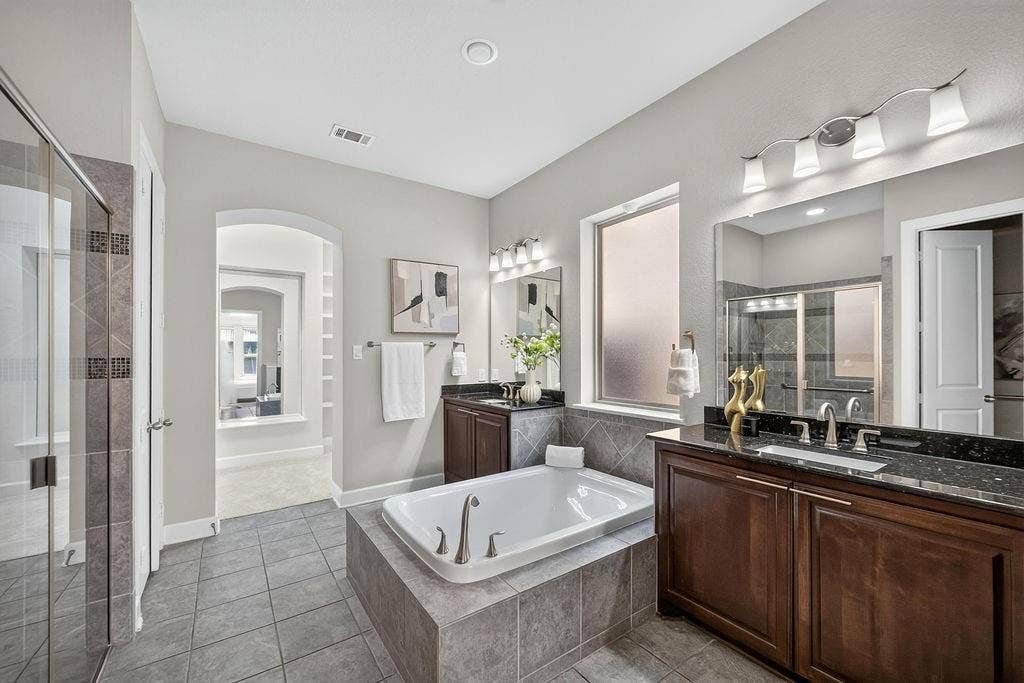 Bathroom featuring a deep soaking tub with tile surround, dark wood cabinetry, dual-sink vanity with dark stone-finish countertops, tiled flooring, and a glass-enclosed walk-in shower