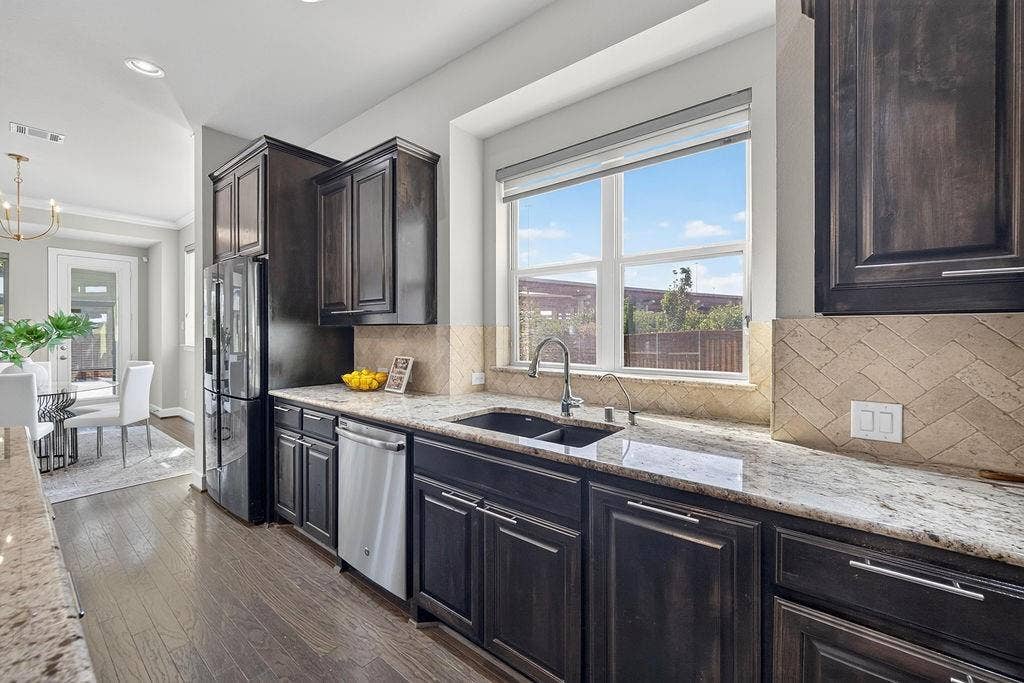Kitchen featuring dark wood cabinetry, natural stone countertops, herringbone-pattern tile backsplash, stainless steel appliances, and wood-finish flooring