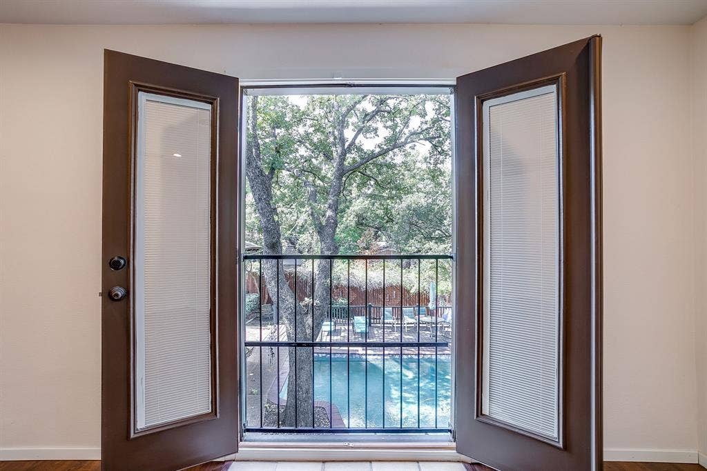 Double doors with integrated blinds opening to a balcony with black metal railing, overlooking a swimming pool and mature trees