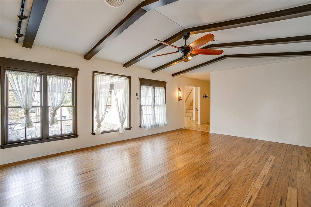 Vaulted ceiling with exposed wood beams, wood-finish flooring, multiple windows, and a ceiling fan with integrated lighting