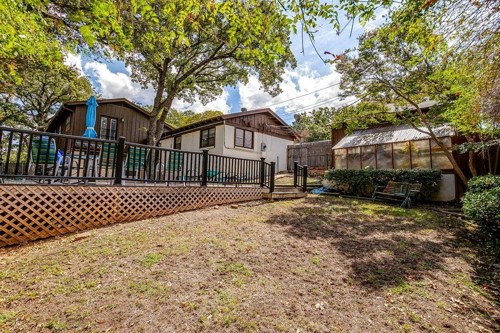 Elevated wood deck with lattice skirting and black railings, leading to a spacious yard with a greenhouse structure and mature trees