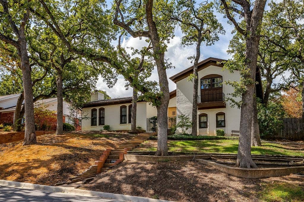 White stucco exterior, brown trim accents, arched window casings, wood-finish balcony, tiered landscaping with mature trees