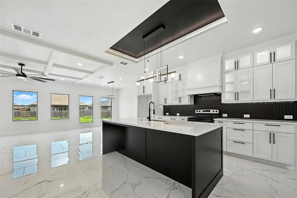 Gleaming white large-format tile flooring, kitchen island with a white countertop and dark cabinetry, white perimeter cabinetry with black hardware, black chevron tile backsplash, and a coffered ceiling with a ceiling fan