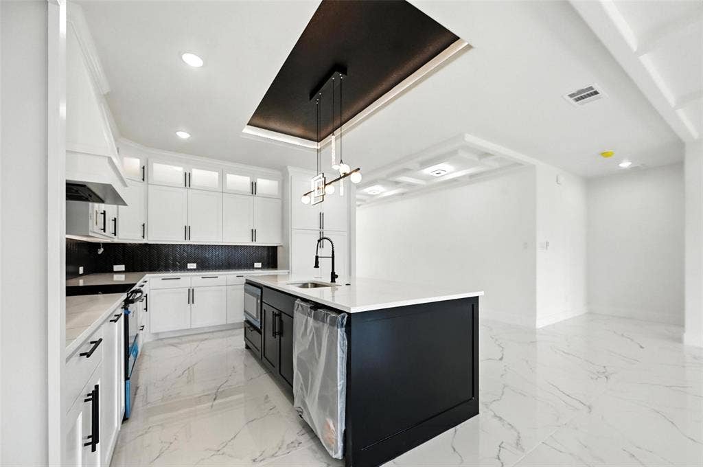 Kitchen featuring white cabinetry, black tile backsplash, central island with sink, and marled tile flooring