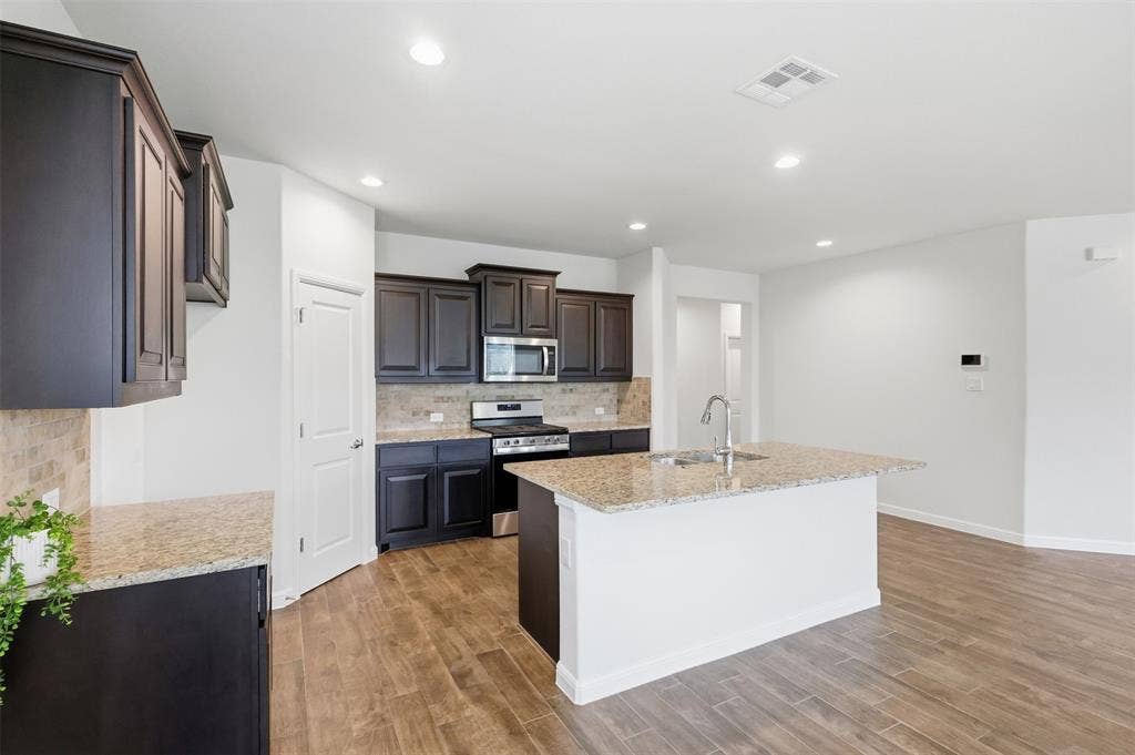 9005 Fescue Drive, Fort Worth, TX 76179 Kitchen featuring dark wood cabinetry, light-toned tiled backsplash, stainless steel appliances, and a central island with a sink and light-toned countertop