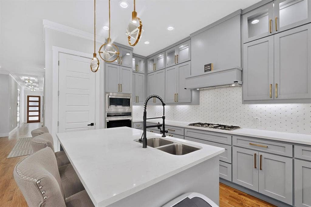 518 Crete Street, Dallas, TX 75203 Kitchen featuring a white countertop island with a double sink, light gray cabinetry with gold-tone hardware, a gas cooktop with an overhead range hood, and wood-finish flooring