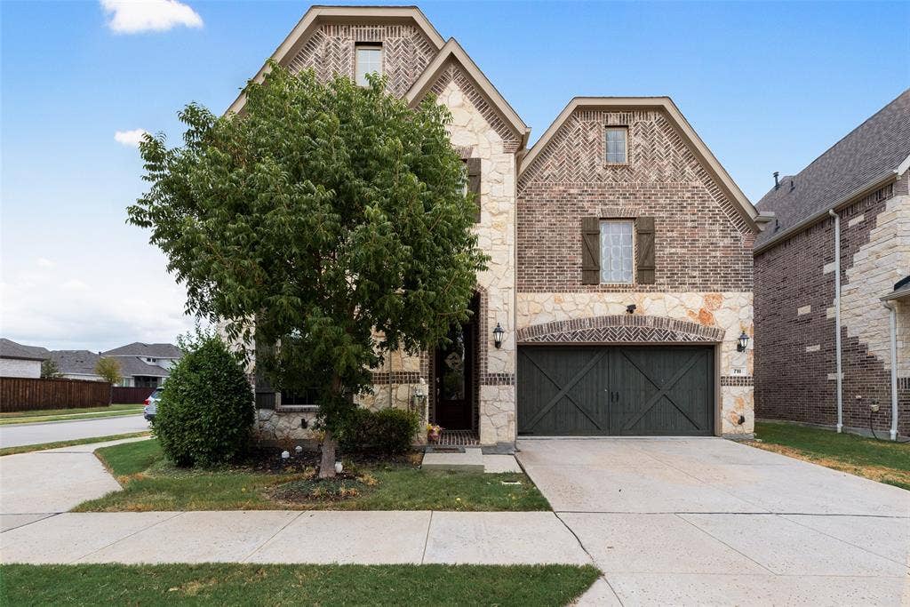 718 Waterbrook Parkway, Argyle, TX 76226 Stone and brick facade with gabled roof lines, featuring an arched garage entrance with a dark wood-finish door, and exterior sconce lighting