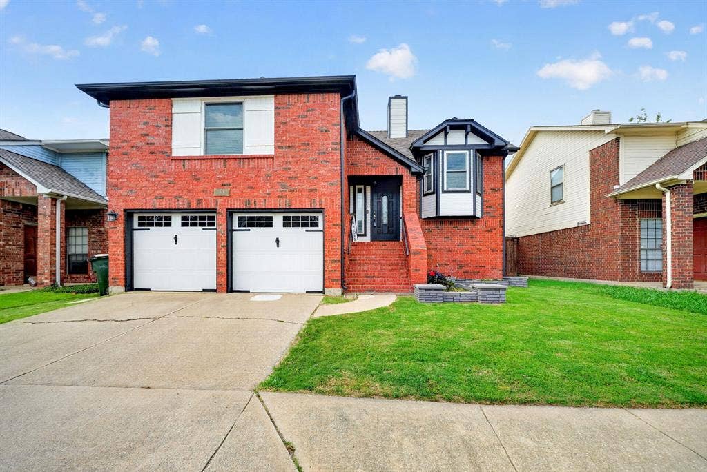 Brick exterior with white window shutters, two white garage doors, a concrete driveway, and a manicured lawn