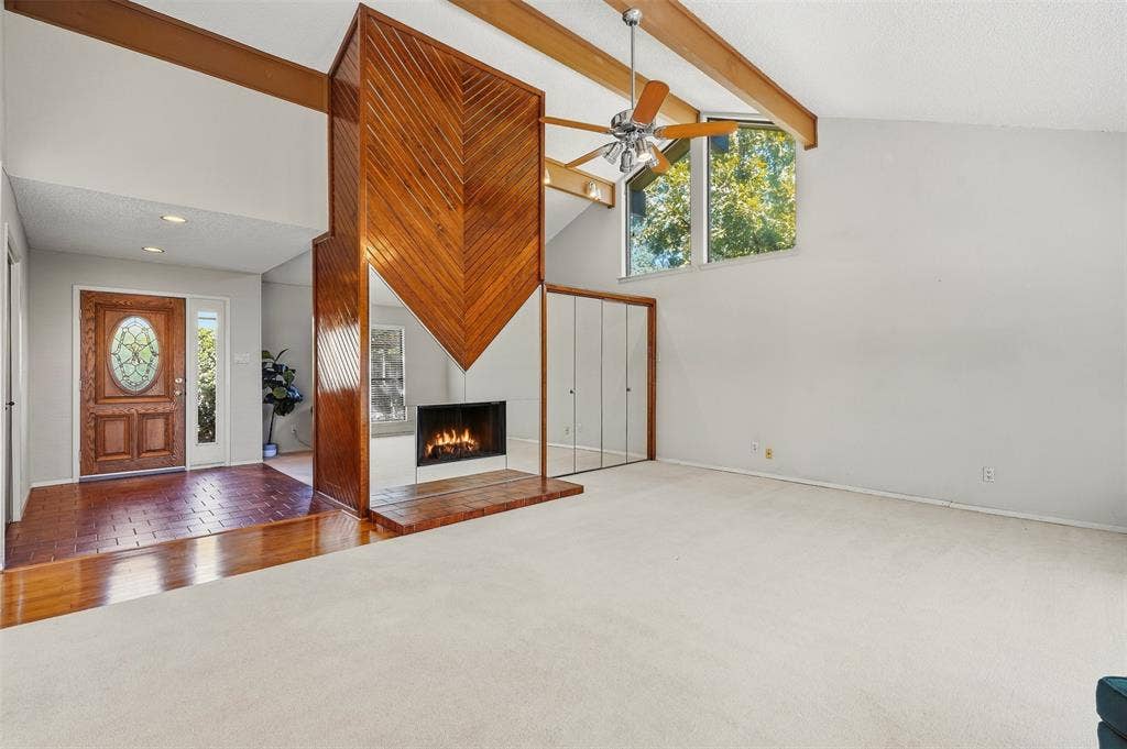 Vaulted ceiling with exposed wood beams, a prominent wood-paneled fireplace, large windows providing natural light, and a wood-paneled entry door with an oval glass insert