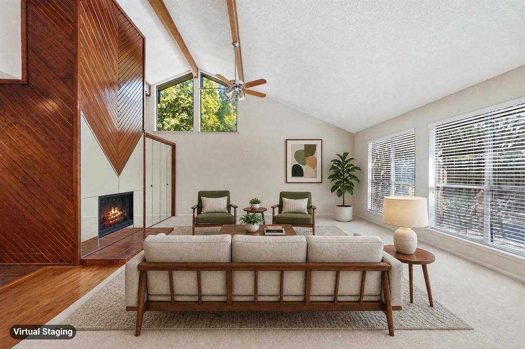 Vaulted ceiling with exposed wood beams, wood-paneled fireplace surround, multiple large windows, and light-colored wall-to-wall carpeting