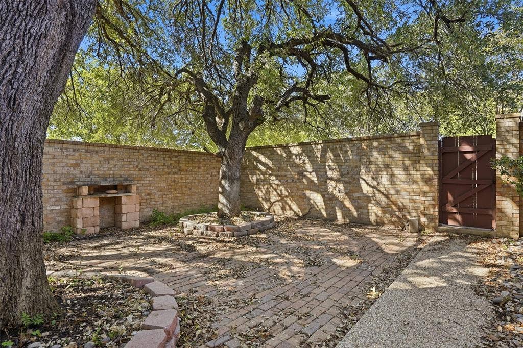 Enclosed brick-walled outdoor area featuring a brick paver pathway, mature trees, a built-in brick structure, and a wooden gate