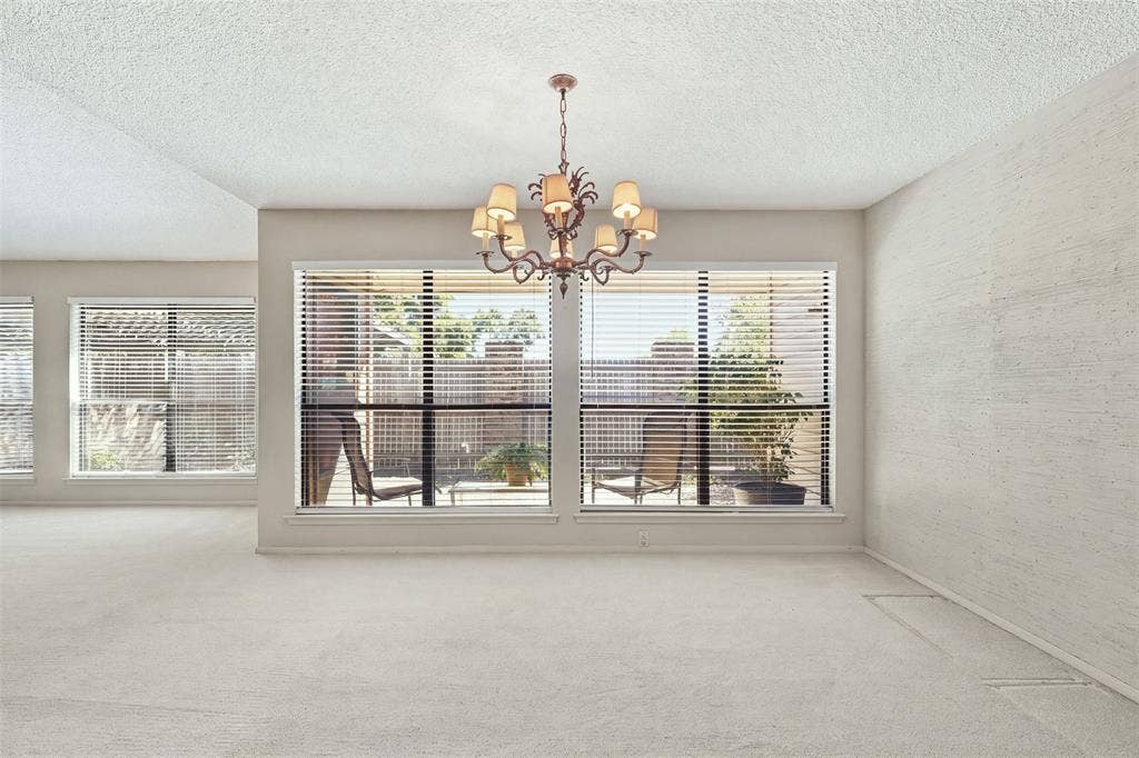 Vaulted ceiling, decorative chandelier, light-colored carpeting, multiple windows with horizontal blinds, and a private outdoor space
