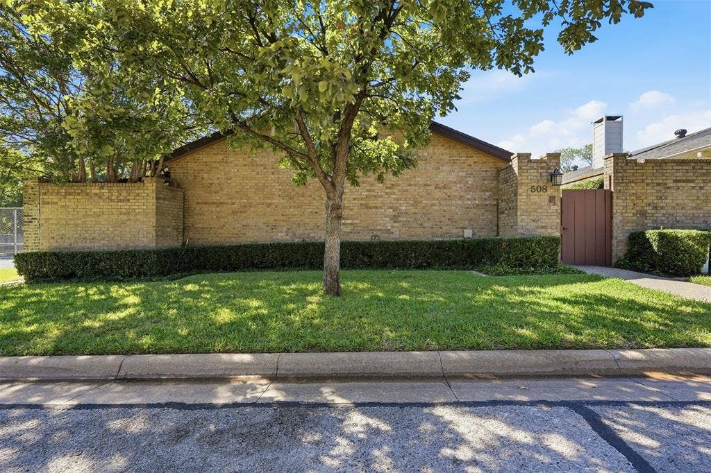 Brick exterior with a peaked roofline, front yard with green lawn, established trees, and a wooden privacy gate