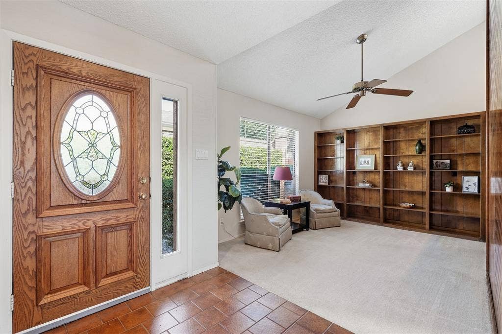 Wood entry door with decorative glass insert, terracotta tile flooring, vaulted ceiling, built-in wood shelving, and ceiling fan