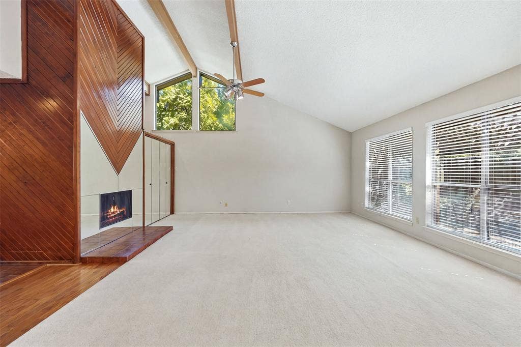 Wood-paneled fireplace surround with reflective paneling, vaulted ceiling with exposed beams, ceiling fan fixture, and multiple windows with blinds
