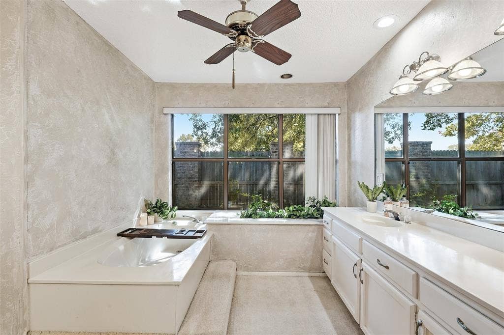Dual-sink vanity with white cabinetry, built-in bathtub, ceiling fan, and large windows